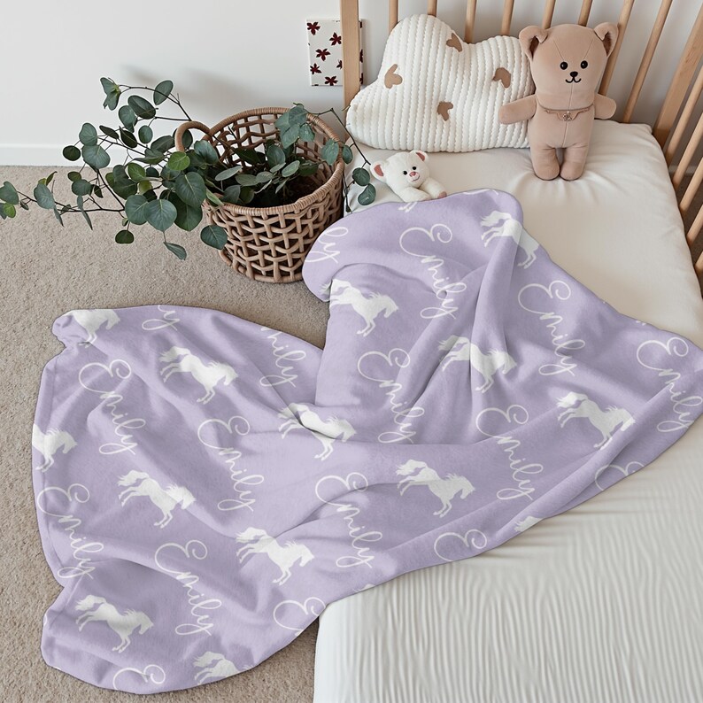 Pu&ograve; includere: Una coperta per neonati lilla con un motivo di cavalli bianchi e il nome "Emily" in corsivo. La coperta &egrave; appoggiata su una culla, con un orsacchiotto e un cuscino a forma di nuvola.