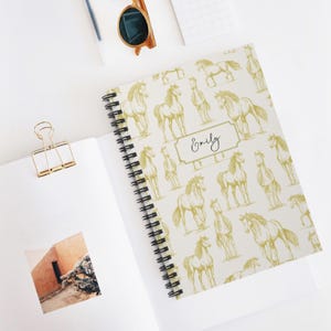 May include: A white spiral notebook with a gold horse pattern and the name "Emily" printed on the cover.
