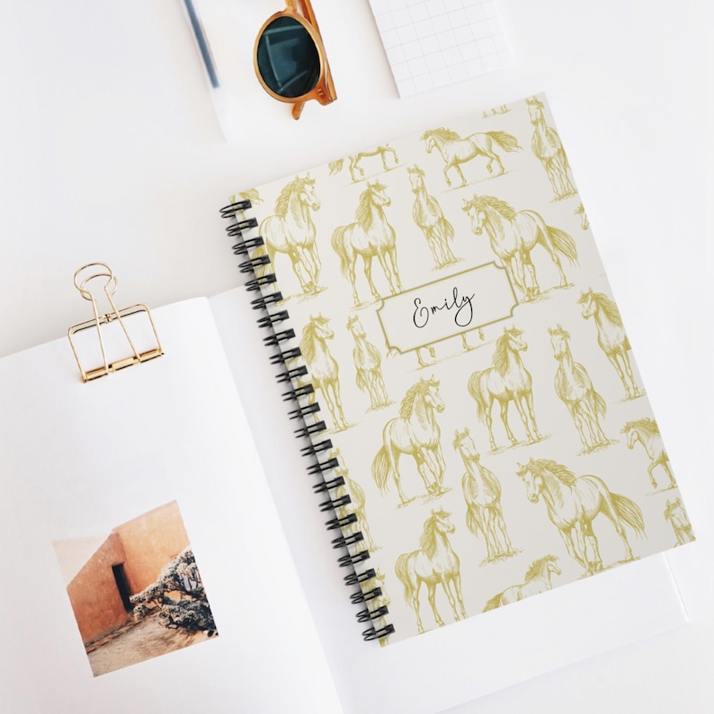 May include: A white spiral notebook with a gold horse pattern and the name "Emily" printed on the cover.