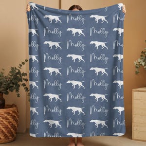 May include: A blue blanket with a repeating pattern of white dog silhouettes and the name "Molly" in a cursive font. The blanket is held up, showcasing the design.