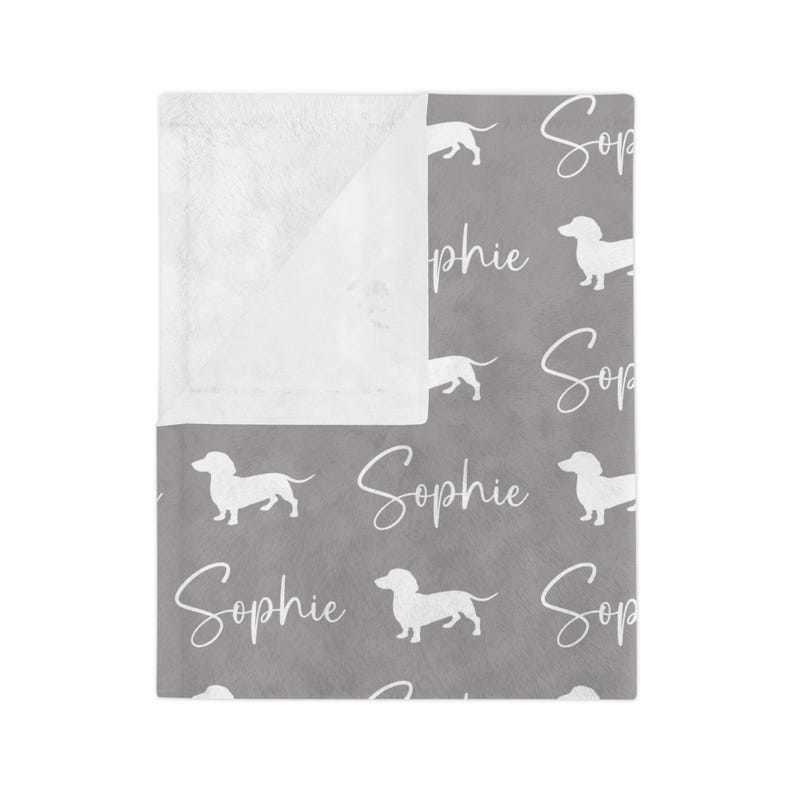 Peut inclure: Une couverture grise orn&eacute;e d'un motif de teckels blancs et du nom "Sophie" en &eacute;criture cursive. La couverture a une bordure blanche et moelleuse. Les teckels sont repr&eacute;sent&eacute;s en position de marche.