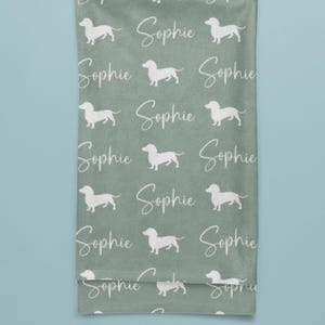 Peut inclure: Une serviette vert sauge avec un motif r&eacute;p&eacute;t&eacute; de silhouettes de chiens teckel blanches et le nom "Sophie" en &eacute;criture cursive. La serviette est pli&eacute;e et suspendue, avec un fond bleu clair.
