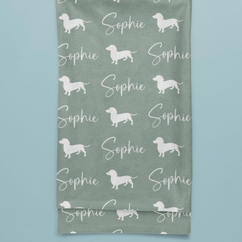 Peut inclure: Une serviette vert sauge avec un motif r&eacute;p&eacute;t&eacute; de silhouettes de chiens teckel blanches et le nom "Sophie" en &eacute;criture cursive. La serviette est pli&eacute;e et suspendue, avec un fond bleu clair.