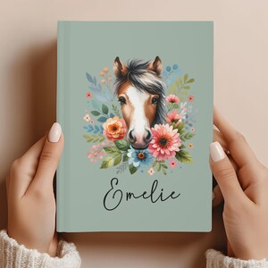 May include: A sage green journal featuring a watercolor illustration of a horse's face surrounded by colorful flowers and foliage. The name "Emelie" is written in cursive below the horse. The journal is held by two hands.