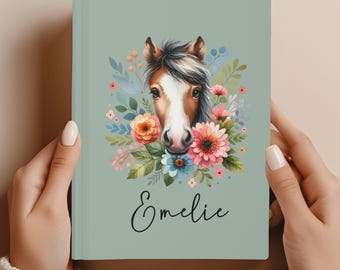 May include: A sage green journal featuring a watercolor illustration of a horse's face surrounded by colorful flowers and foliage. The name "Emelie" is written in cursive below the horse. The journal is held by two hands.
