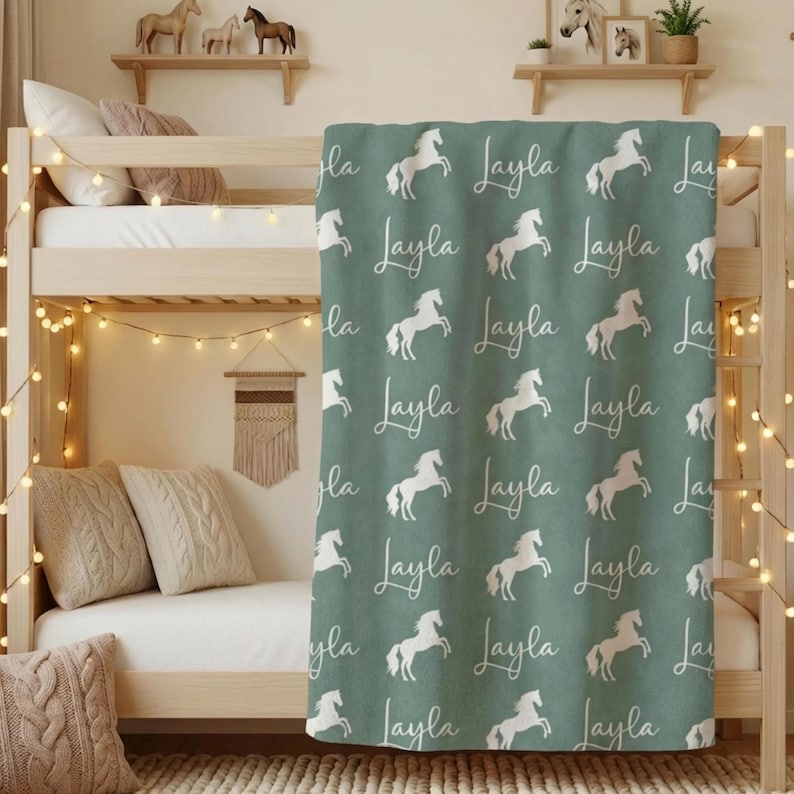 Pu&ograve; includere: Una coperta verde con un motivo ripetuto di cavalli bianchi e il nome "Layla" in corsivo. La coperta &egrave; drappeggiata su un letto a castello, con luci decorative e cuscini visibili.