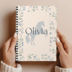 May include: A spiral-bound notebook with a cream-colored cover featuring a light blue horse silhouette and the name "Olivia" in gray. The cover is framed with a floral pattern in blue and green.