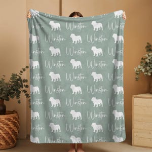 Person holding up a large custom English Bulldog blanket showing the repeating pattern of english bulldog silhouettes and the custom name in white on an aqua green background.