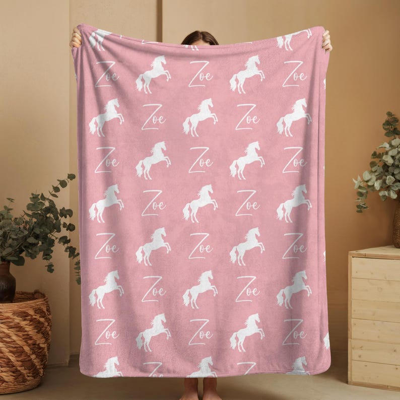 Pu&ograve; includere: Una coperta rosa con un motivo ripetuto di sagome di cavalli bianchi e il nome "Zoe" in scrittura bianca. La coperta &egrave; esposta, evidenziando il suo design e il colore.