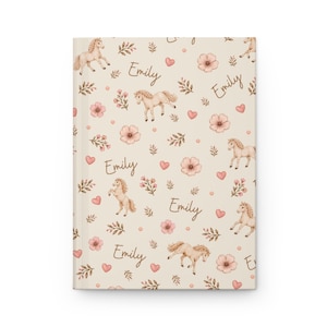 May include: A hardcover journal with a cream-colored background featuring a repeating pattern of tan horses, pink flowers, hearts, and the name "Emily" in a script font. The journal is closed and has a clean, crisp appearance.