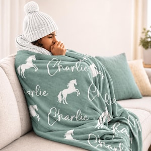 Pu&ograve; includere: Una coperta color verde acqua con silhouette di cavalli bianchi e il nome "Charlie" in corsivo. La coperta &egrave; drappeggiata su una persona che indossa un cappello e una sciarpa di lana bianchi. Un cuscino verde acqua &egrave; visibile.