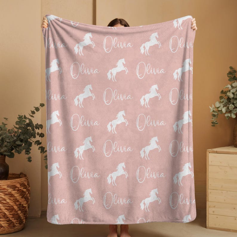 Pu&ograve; includere: Una coperta rosa con un motivo ripetuto di cavalli bianchi rampanti e il nome "Olivia" in corsivo. La coperta &egrave; morbida e sembra essere realizzata in un materiale felpato.