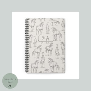 May include: A white spiral bound notebook with a gray horse pattern and the name "Emily" written on the front.
