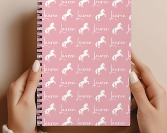 May include: A spiral-bound notebook with a pink cover featuring a repeating pattern of white rearing horses and the name "Lauren" in a cursive font. The notebook is held open, displaying the design.