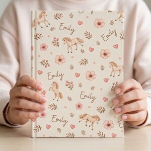 May include: A cream-colored journal featuring a repeating pattern of tan horses, pink flowers, and hearts. The name "Emily" is written in a script font throughout the design. The journal is held open, showcasing the cover's design.
