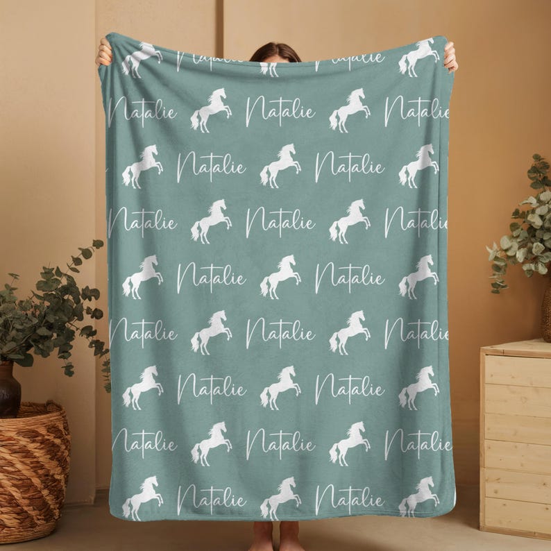 Pu&ograve; includere: Una coperta turchese con un motivo ripetuto di cavalli bianchi rampanti e il nome "Natalie" in corsivo. La coperta &egrave; esposta, evidenziando il suo design e il suo colore.