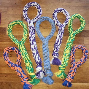 May include: A collection of colorful dog rope toys in various shades of blue, purple, green, yellow, and orange. Each toy features a braided rope design with a loop at one end and a knotted tassel at the other. The toys are arranged on a wooden surface.