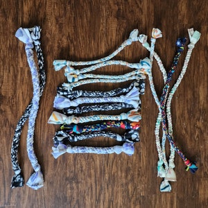 May include: A collection of colorful, patterned dog toys made from fabric rope. The toys are tied together in a bundle and feature various colors and patterns, including blue, white, black, and red.