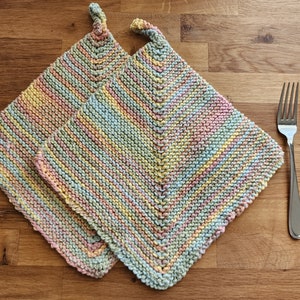 May include: Two knitted pot holders in a multi-colored striped pattern of pink, yellow, blue, and green. The pot holders are folded on top of each other on a wooden surface.