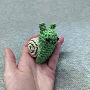 Puede incluir: Un caracol de ganchillo hecho a mano en verde y crema. El cuerpo del caracol es verde, con una concha en espiral en crema y marrón. Tiene ojos negros y pequeñas antenas. El caracol se sostiene en una mano.