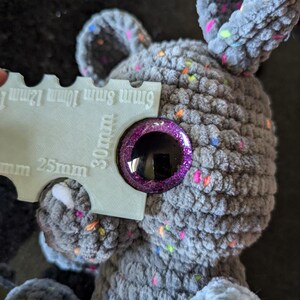 Peut inclure: Gros plan sur un animal en peluche gris crochet&eacute; avec un &oelig;il violet et paillet&eacute;. Une r&egrave;gle est utilis&eacute;e pour montrer que l'&oelig;il a un diam&egrave;tre de 30 mm.