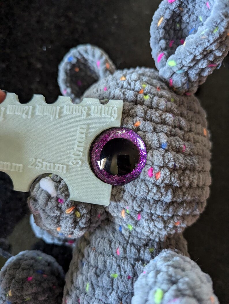 Peut inclure: Gros plan sur un animal en peluche gris crochet&eacute; avec un &oelig;il violet et paillet&eacute;. Une r&egrave;gle est utilis&eacute;e pour montrer que l'&oelig;il a un diam&egrave;tre de 30 mm.