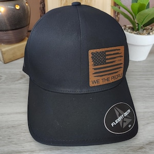 Könnte beinhalten: Schwarze Baseballkappe mit gebogenem Schirm. Ein brauner Lederaufnäher zeigt ein amerikanisches Flaggen-Design und den Text "WE THE PEOPLE". Das Flexfit Delta-Logo befindet sich auf dem Schirm.