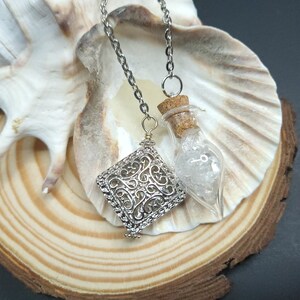 May include: Two silver chains with charms hanging from them. One charm is a square filigree design, the other is a small glass bottle with a cork stopper filled with white crystals.