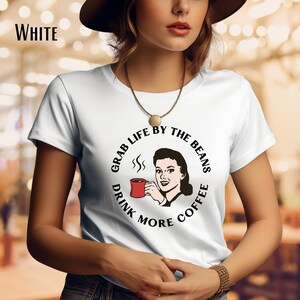May include: White t-shirt featuring a graphic of a woman holding a cup of coffee and the text "Grab Life By The Beans Drink More Coffee".