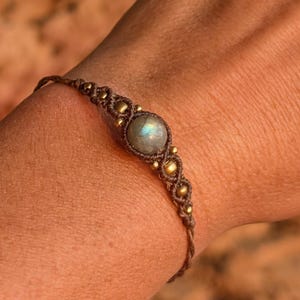 Può includere: Un braccialetto in macramè marrone con una gemma centrale rotonda blu chiaro. Il braccialetto è adornato con piccole perline color oro ed è mostrato sul braccio di una persona.