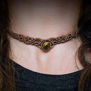 May include: A brown macrame choker necklace featuring a central round tiger's eye gemstone. The cord is intricately knotted in a repeating pattern, with small metallic beads flanking the stone on both sides to create a detailed, textured design.