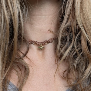 Könnte beinhalten: Ein braunes Makramee-Choker-Halskette mit einem goldfarbenen Bienenanhänger. Die Halskette wird um den Hals getragen. Der Hintergrund ist verschwommen, mit langen, welligen blonden Haaren und einem grauen T-Shirt.