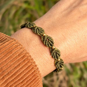 May include: A green friendship bracelet with gold beads, woven in a leaf pattern.