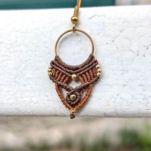 Puede incluir: Un pendiente de macramé con un aro y un gancho de metal dorado. El diseño incorpora cordón anudado en marrón oscuro y marrón claro dispuesto en un patrón triangular, acentuado con pequeñas cuentas de metal dorado integradas en la estructura.