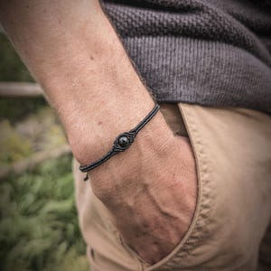 Op de afbeelding: Zwart macramé armband met een centrale, ronde, zwarte steen. De armband wordt om de pols gedragen. De persoon draagt een grijze trui en een kaki broek. Een eenvoudig, elegant sieraad.