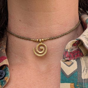Op de afbeelding: Een goudkleurige spiraalvormige hangerketting. De hanger hangt aan een geweven, olijfgroene koordketting. De ketting wordt gedragen tegen de huid van een persoon, met een patroonshirt zichtbaar op de achtergrond.