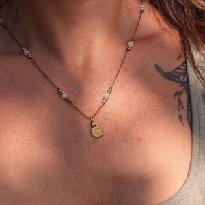 Op de afbeelding: Een delicate goudkleurige ketting met kleine, ronde kralen en een hanger. De ketting heeft een fijne ketting met gelijkmatig verdeelde kralen en een kleine, cirkelvormige hanger met een gedetailleerd zonnepatroon. De ketting wordt door een persoon gedragen.