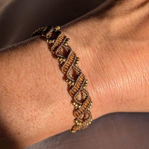May include: A brown and gold braided bracelet with small gold beads. The bracelet is woven in a diamond pattern.