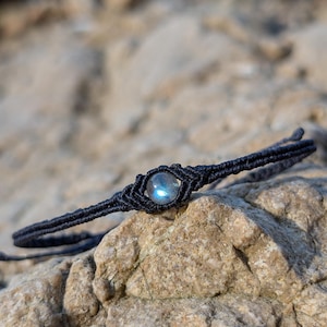 Peut inclure: Un bracelet macramé noir avec une pierre précieuse centrale, ronde et irisée. Le bracelet est présenté sur une roche texturée brun clair. Le macramé est complexe, et la pierre précieuse a un léger scintillement bleu et vert.