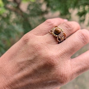 Puede incluir: Un anillo de macramé hecho a mano con una piedra preciosa central redonda y marrón. El anillo tiene un diseño tejido en tonos beige y marrón, con pequeñas cuentas doradas que acentúan el engaste de la piedra preciosa. El anillo se muestra en una mano.