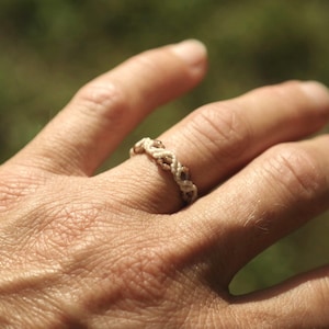 Può includere: Primo piano di una mano che indossa un anello intrecciato bianco e marrone. L'anello è realizzato con un materiale intrecciato e ha un design semplice.