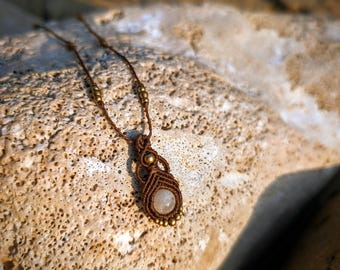 May include: A brown macrame necklace featuring a central round, translucent stone set within intricate knotted cord patterns. The design includes small metallic beads integrated into the cord and along the lower edge of the pendant setting.