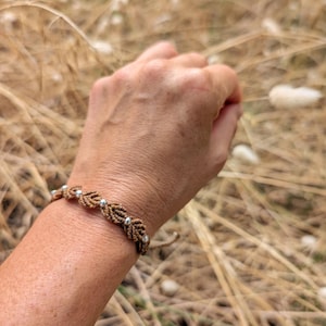 May include: A macrame bracelet with a leaf design and silver beads. The bracelet is made of brown cord and is worn on a wrist. The background is blurred, showing dry grass and a natural setting.