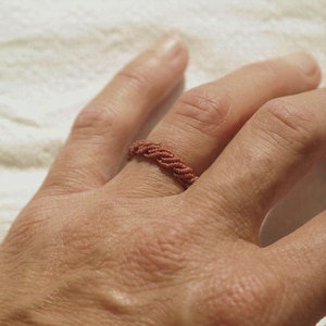 May include: A ring crafted from twisted terracotta-colored cord, featuring a braided texture that wraps around the finger. The band is constructed from multiple strands of thin, durable thread woven together to create a consistent, repeating pattern.