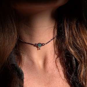 Peut inclure: Un collier ras-du-cou en macramé marron foncé avec une pierre précieuse ronde verte au centre, de petites perles métalliques dorées et une petite breloque dorée suspendue. Le cordon est fin et tissé avec un motif de nœuds texturé.