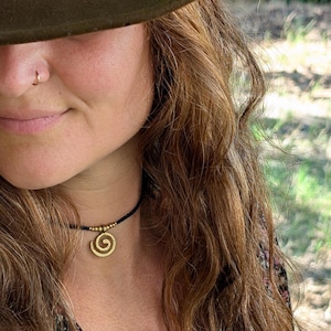 May include: A gold spiral pendant necklace on a black cord. The pendant features a stylized spiral design. The necklace is worn against a backdrop of long, wavy brown hair and a dark floral print top. A green hat is partially visible.
