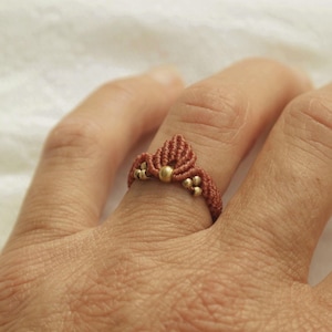 Puede incluir: Un anillo de cordón marrón con cuentas doradas. El anillo se lleva en un dedo.