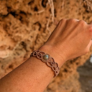 May include: A macrame bracelet in shades of brown, featuring a central stone and gold-colored beads. The woven design is intricate, with a central stone. The bracelet is worn on a wrist, showcasing the earthy tones and craftsmanship.