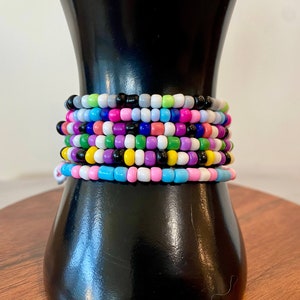 May include: A stack of colorful beaded bracelets in shades of pink, blue, green, yellow, purple, and black. The bracelets are arranged on a black mannequin.