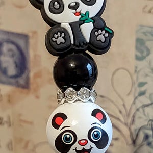 May include: A black pen with a panda bear charm. The charm is white with black eyes and a pink bow. The pen is decorated with black beads and a silver rhinestone band.